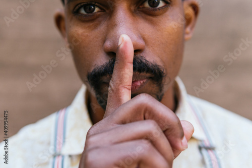 Crop African American male covering mouth with finger and showing silence sign while looking at camera photo