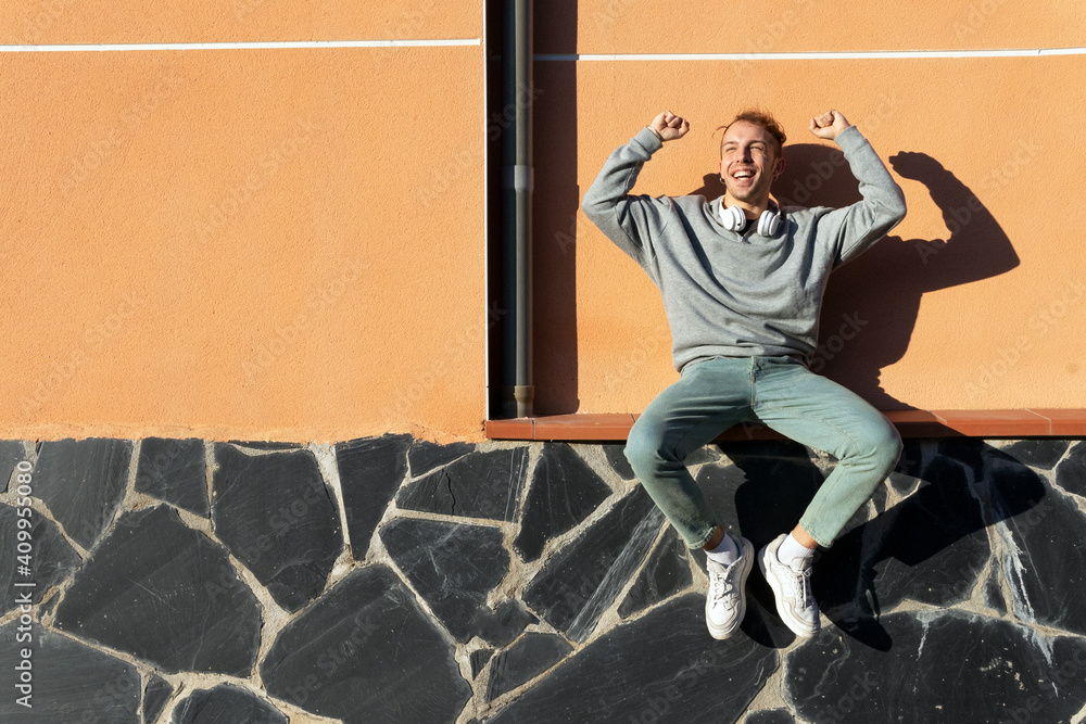 Low angle full body of delighted energetic young hipster male in casual clothes with headphones on neck sitting on bench near wall and raising arms while rejoicing over good news on street Low angle full body of delighted energetic young hipster male in casual clothes with headphones on neck sitting on bench near wall and raising arms while rejoicing over good news on street