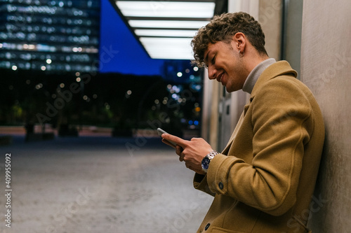 Side view of smiling handsome male leaning on building and messaging on social media via smartphone in evening in city