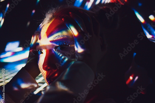 Asian female model with creative makeup standing in studio illuminated by colorful projector lights and looking at camera while touching head photo