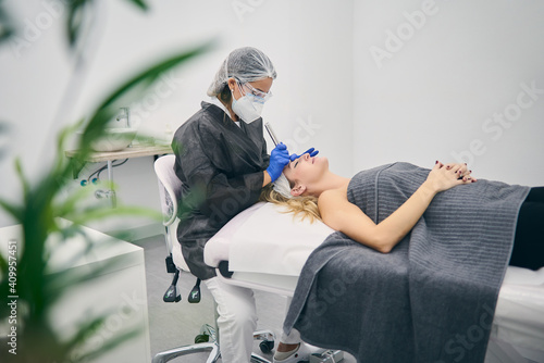 Side view of cosmetologist in uniform using derma pen with micro needles and cleaning face of client in beauty clinic photo