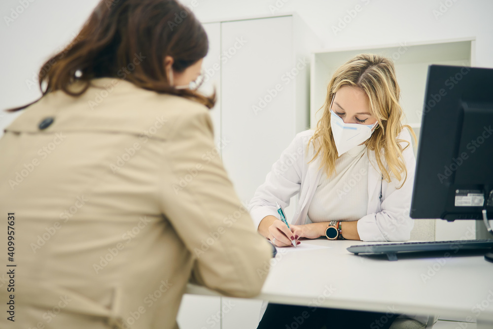 Female dermatologist with face mask sitting at table with client and taking notes on paper while writing prescription for buying cosmetic products during coronavirus pandemic Female dermatologist with face mask sitting at table with client and taking notes on paper while writing prescription for buying cosmetic products during coronavirus pandemic