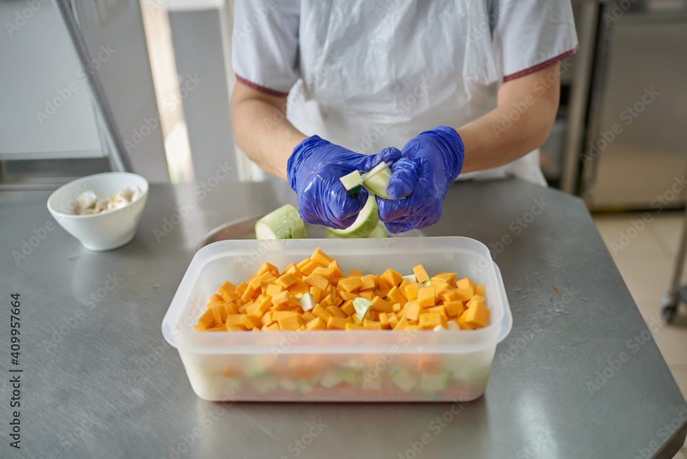 Female cook in white uniform and protective mask and gloves cutting fresh vegetables while preparing food in hospital kitchen during coronavirus pandemic Female cook in white uniform and protective mask and gloves cutting fresh vegetables while preparing food in hospital kitchen during coronavirus pandemic