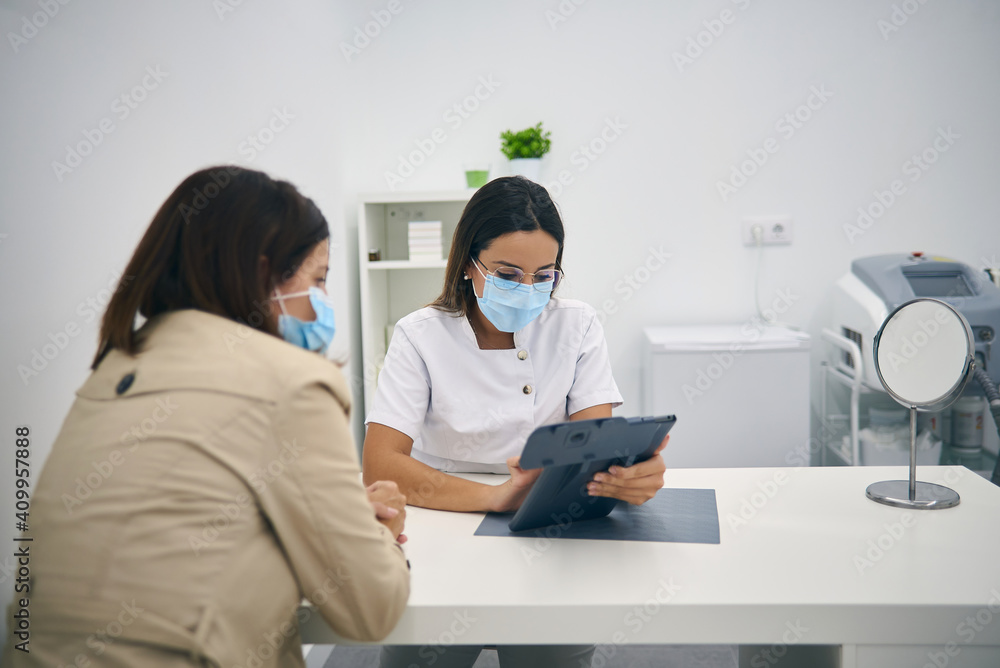 Female client in mask sitting in modern beauty clinic and discussing skin care procedures with cosmetologist during appointment Female client in mask sitting in modern beauty clinic and discussing skin care procedures with cosmetologist during appointment