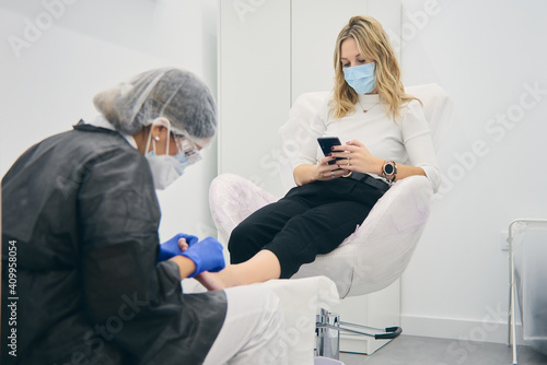 Female podologist in uniform doing pedicure for woman sitting in medical chair in beauty clinic photo