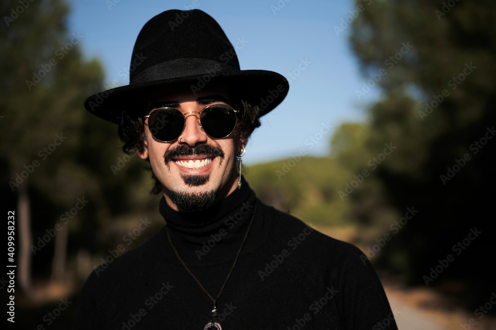 Side view of cheerful young hipster bearded male traveler with piercing wearing stylish black hat and sunglasses looking at camera while standing on blurred country road Side view of cheerful young hipster bearded male traveler with piercing wearing stylish black hat and sunglasses looking at camera while standing on blurred country road