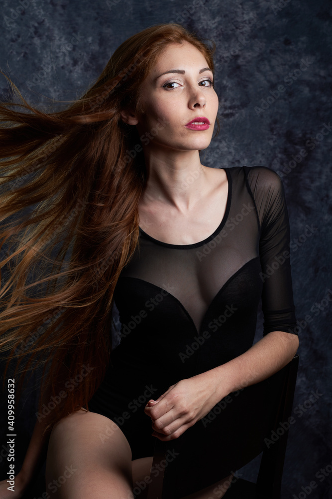 Calm female model in stylish clothes sitting in chair in studio with red flying hair and looking at camera Calm female model in stylish clothes sitting in chair in studio with red flying hair and looking at camera