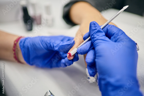 Crop master of manicure using metal spatula and removing gel polish from nails of client in beauty salon photo