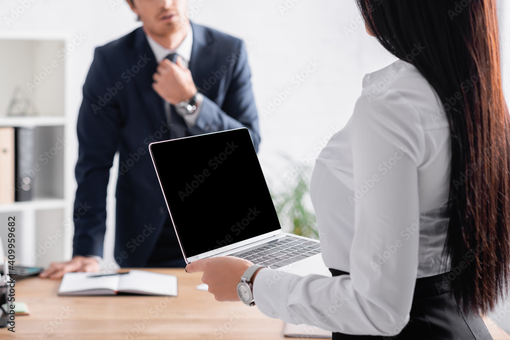 cropped view of sexy businesswoman holding laptop with blank screen near colleague adjusting tie on blurred background cropped view of sexy businesswoman holding laptop with blank screen near colleague adjusting tie on blurred background