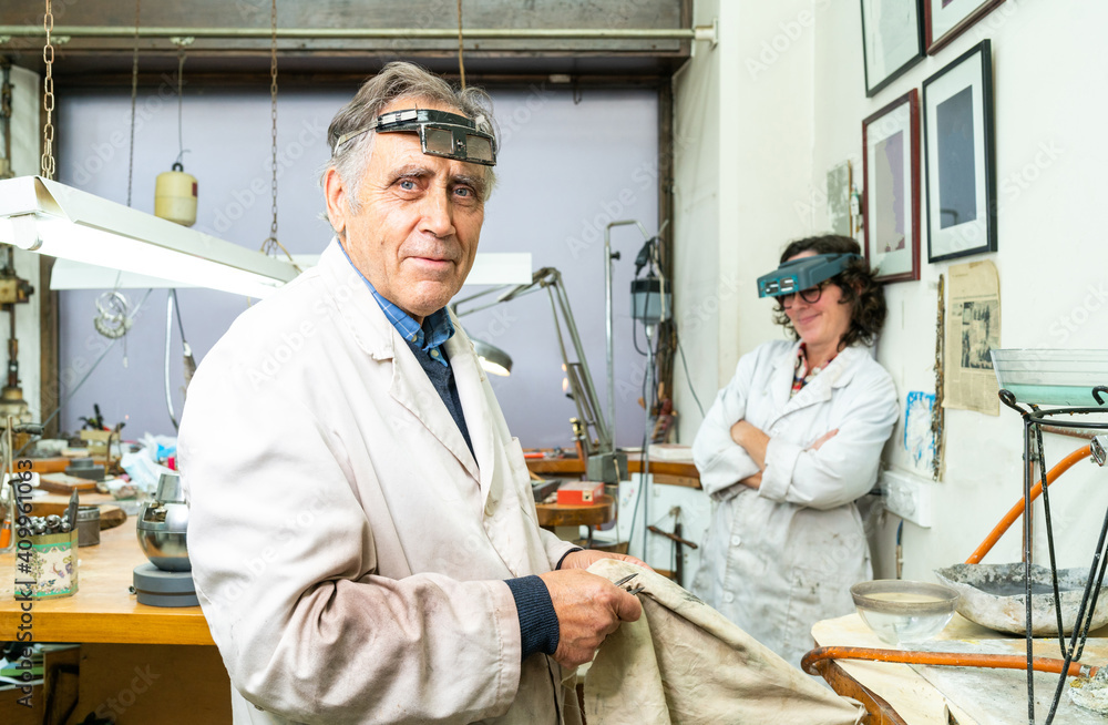 Side view of mature gray haired professional male jeweler in work wear and with glasses on forehead looking at camera while working with female colleague in workshop Side view of mature gray haired professional male jeweler in work wear and with glasses on forehead looking at camera while working with female colleague in workshop