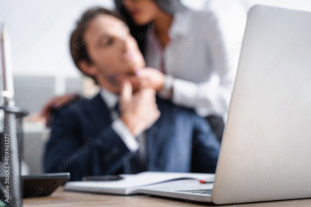 selective focus of laptop near businesswoman flirting with young colleague on blurred background selective focus of laptop near businesswoman flirting with young colleague on blurred background