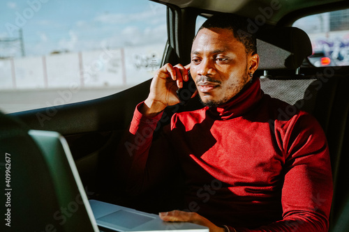 Busy African American businessman sitting in luxury car with laptop and speaking with client on smartphone while looking away photo