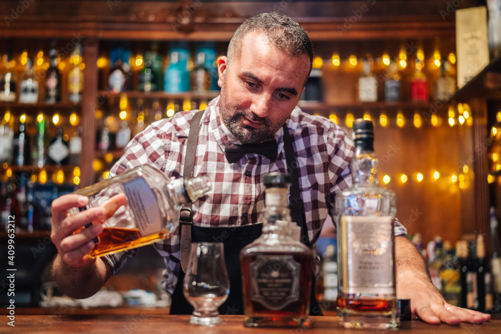 Crop brutal male bartender in apron pouring brandy in glass placed on counter with bottles of alcoholic drinks Crop brutal male bartender in apron pouring brandy in glass placed on counter with bottles of alcoholic drinks