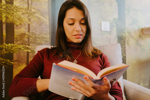 Concentrated female sitting in armchair and enjoying fascinating story in book while relaxing at weekend at home