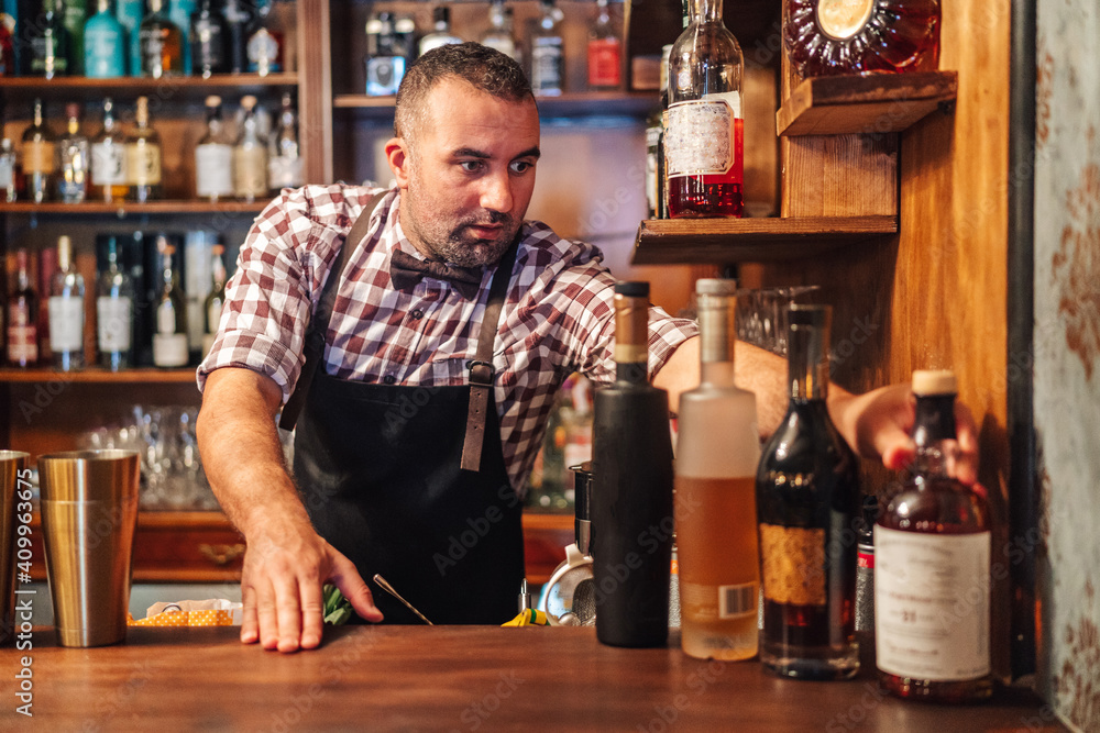 Crop brutal male bartender in apron grabbing bottle with alcoholic drinks placed on counter Crop brutal male bartender in apron grabbing bottle with alcoholic drinks placed on counter