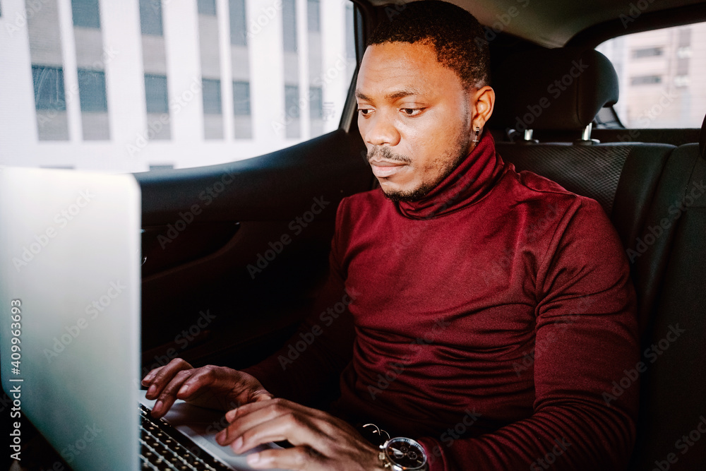 Focused African American male entrepreneur sitting in car on backseat and browsing netbook while working remotely on project Focused African American male entrepreneur sitting in car on backseat and browsing netbook while working remotely on project