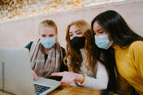 Group of multiethnic female freelancers in masks working on laptop in cafe while discussing new business project during coronavirus epidemic photo