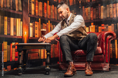 Full body of man in elegant suit holding glass of Irish coffee while resting in comfortable armchair photo