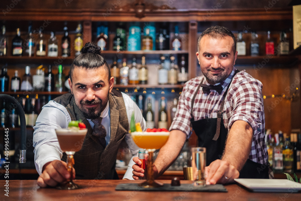 Bearded bartenders smiling while holding glasses of fresh cold sour cocktails at counter in bar Bearded bartenders smiling while holding glasses of fresh cold sour cocktails at counter in bar