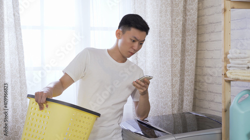 asian man leaning on washing machine with hand holding a basket, heard a strange sound and checking to see if the appliance on cellphone runs smoothly.