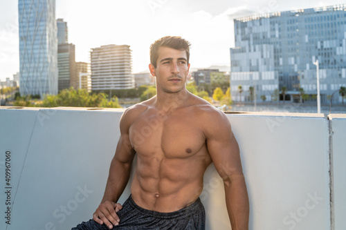 Muscular shirtless male standing near stone fence resting after fitness training in city in summer day looking away