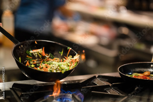 Skillet pan with fried vegetables being stirred by anonymous cook during preparation on gas stove in restaurant kitchen