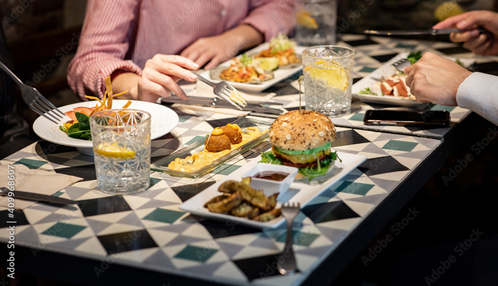 Group of crop anonymous friends gathering at table with various snacks and drinks while having dinner together in cozy restaurant Group of crop anonymous friends gathering at table with various snacks and drinks while having dinner together in cozy restaurant