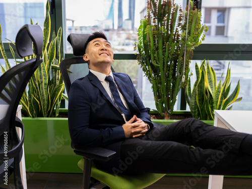 Side view of cheerful young Asian businessman in formal suit looking up and smiling dreamfully while thinking about successful business strategy in modern workspace photo