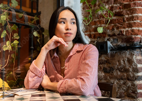 Young tranquil Asian female looking away in casual clothes sitting alone at table in cafe and waiting for order photo