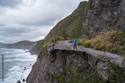 Unrecognizable distant male traveler standing on edge of destroyed asphalt road running along rough rocky coast near waving sea in overcast weather photo