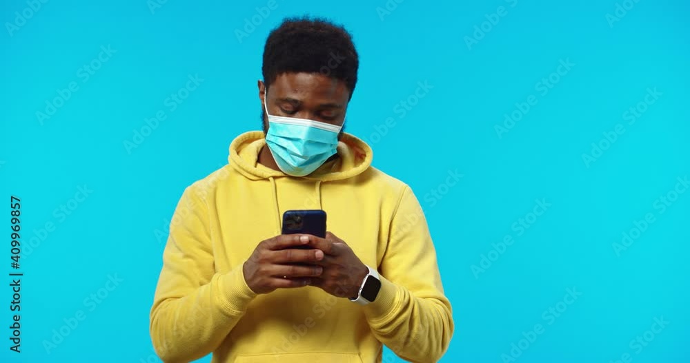 Close up portrait of African American handsome young guy in medical mask texting on smartphone isolated on blue background, browsing social network app on mobile phone, technology communication Close up portrait of African American handsome young guy in medical mask texting on smartphone isolated on blue background, browsing social network app on mobile phone, technology communication