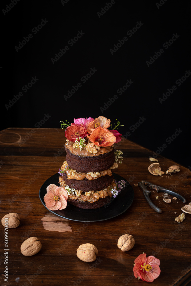 Delectable chocolate naked cake with cream and walnuts garnished with fresh flowers served on wooden table Delectable chocolate naked cake with cream and walnuts garnished with fresh flowers served on wooden table