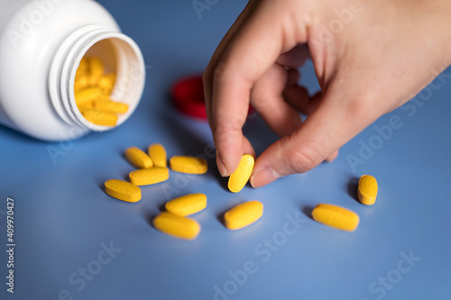 Closeup crop hand of unrecognizable female taking yellow vitamin pills scattered from plastic bottle on blue table photo