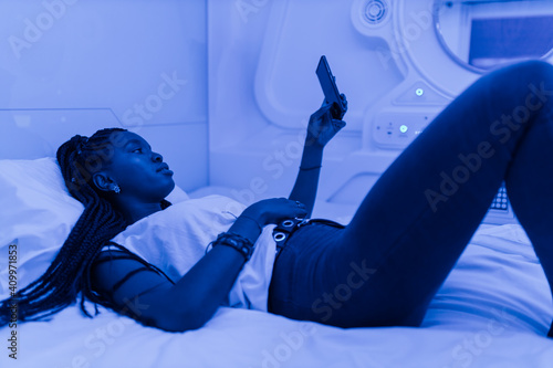 Side view of young African American female tourist lying down on bed and browsing on mobile phone while spending night in contemporary capsule hotel photo