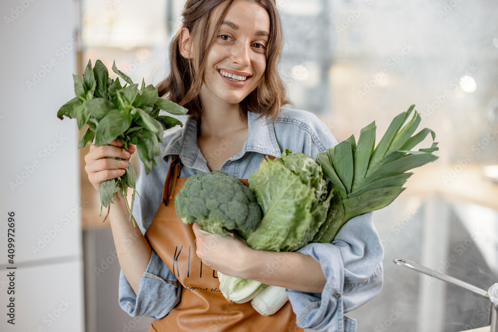 Obraz premium Portrait of pretty smiling woman with fresh broccoli, roman salad, basil on the kitchen with steam on background. Healthy green vegetable concept. Close up. High quality photo