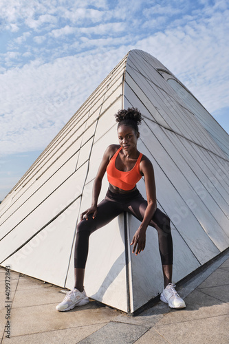 Confident African American sportswoman with slim body sitting on geometric construction in city and looking at camera photo