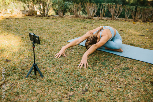 Female blogger recording video on smartphone for social media while practicing yoga on mat in Baddha Konasana in summer park photo