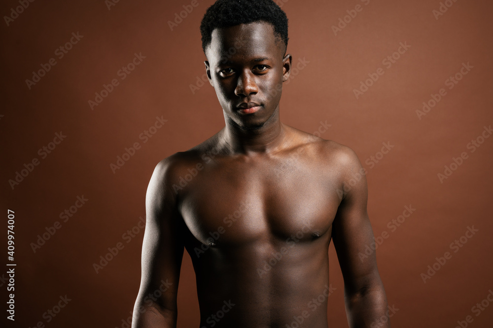Confident African American young male with muscular naked torso standing in trendy jeans with hand on waist on brown background in studio and looking at camera Confident African American young male with muscular naked torso standing in trendy jeans with hand on waist on brown background in studio and looking at camera