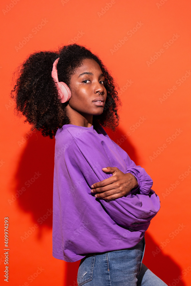 Modern unemotional black woman with Afro hairstyle wearing pink headphones and listening to music on red background Modern unemotional black woman with Afro hairstyle wearing pink headphones and listening to music on red background