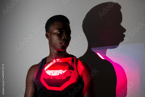 Emotionless African American male model standing in studio with red neon light projection of stop road sign showing concept of restriction photo
