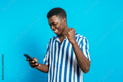 Delighted African American male browsing smartphone and reading good news about business project while standing with clenched fist on vivid blue background in studio