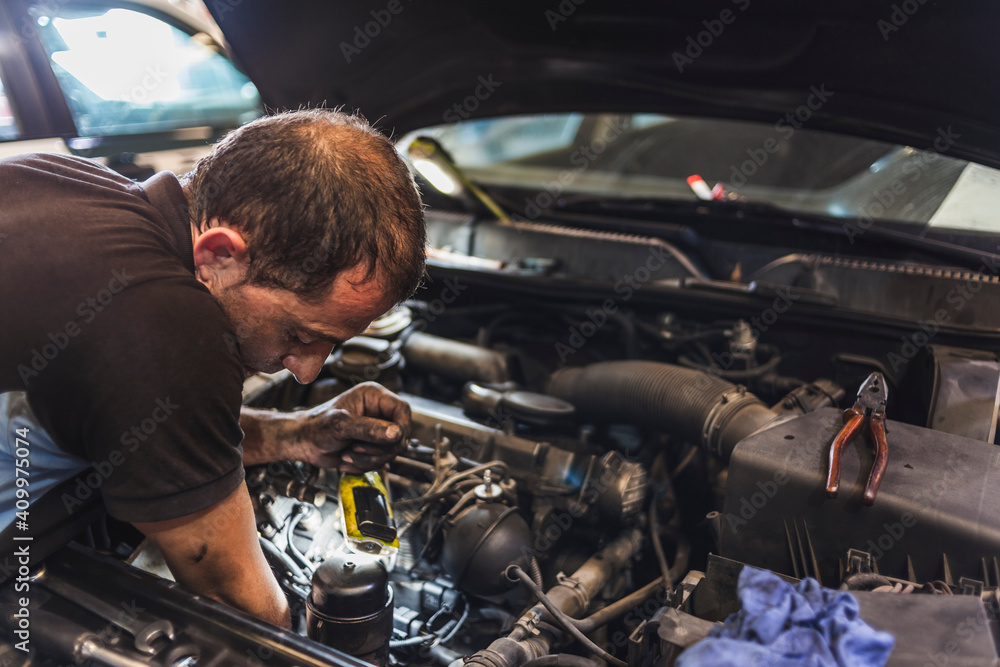 Side view of adult male mechanic illuminating engine with flashlight and checking motor of vehicle during work in garage Side view of adult male mechanic illuminating engine with flashlight and checking motor of vehicle during work in garage