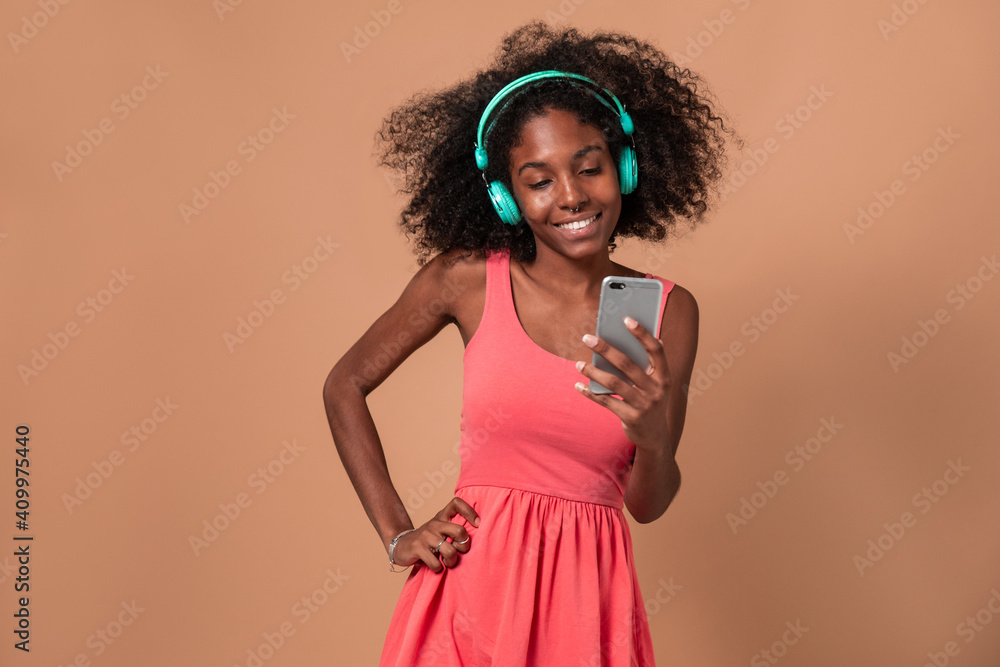 Casual content black woman with Afro hairstyle using smartphone while listening to music with headphones on brown background Casual content black woman with Afro hairstyle using smartphone while listening to music with headphones on brown background
