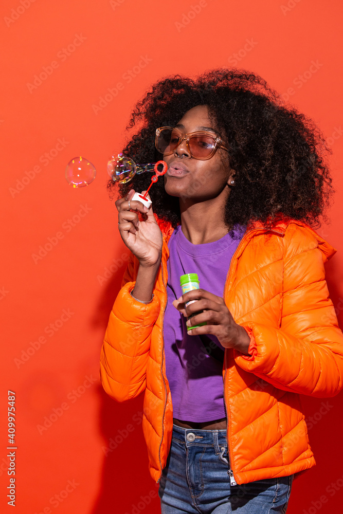 Modern African American female in colorful jacket blowing soap bubbles having fun in studio on red background Modern African American female in colorful jacket blowing soap bubbles having fun in studio on red background