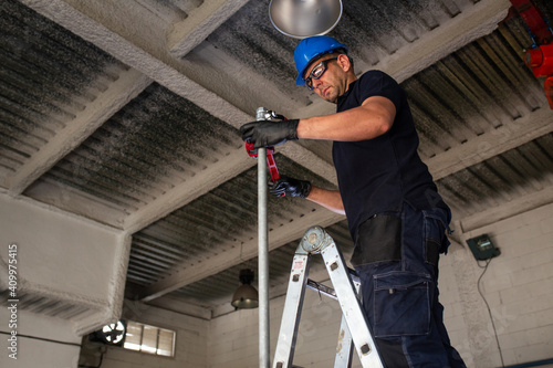 Low angle of cheerful male worker in hardhat and protective glasses standing on ladder and installing metal pipe with plumber wrench while working in workshop