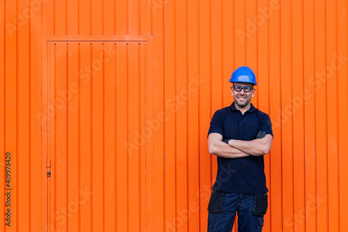 Positive confident adult male workman in blue protective hardhat and eyeglasses smiling friendly while standing against bright orange wall of workshop photo