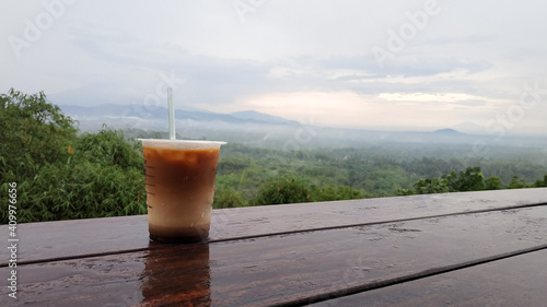 A plastic cup filled with iced coffee on the table with a backdrop of mountain views in the Manoreh Hills area, Central Java - Indonesia  photo