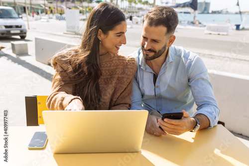 Romantic young ethnic guy in classic outfit hugging and kissing cheek of stylish girlfriend working distantly on laptop in street cafe near sea photo