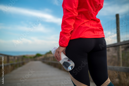 Cropped unrecognizable female athlete holding plastic bottle with fresh water standing on wooden promenade photo