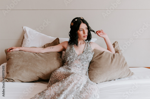 Calm female in sparkling elegant dress resting on comfortable bed with pillows while touching hair and looking away photo